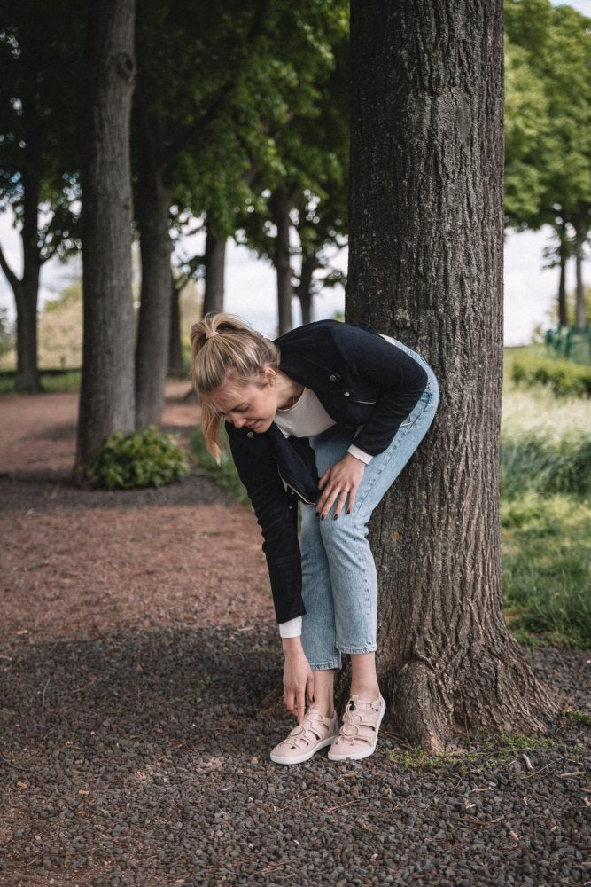 Een vrouw met blond haar, gekleed in een zwarte jas, een wit overhemd en jeans, bukt zich om haar QLEAR Light Pink van ZAQQ te corrigeren, terwijl ze op een met bomen omzoomd grindpad tegen een boom leunt.