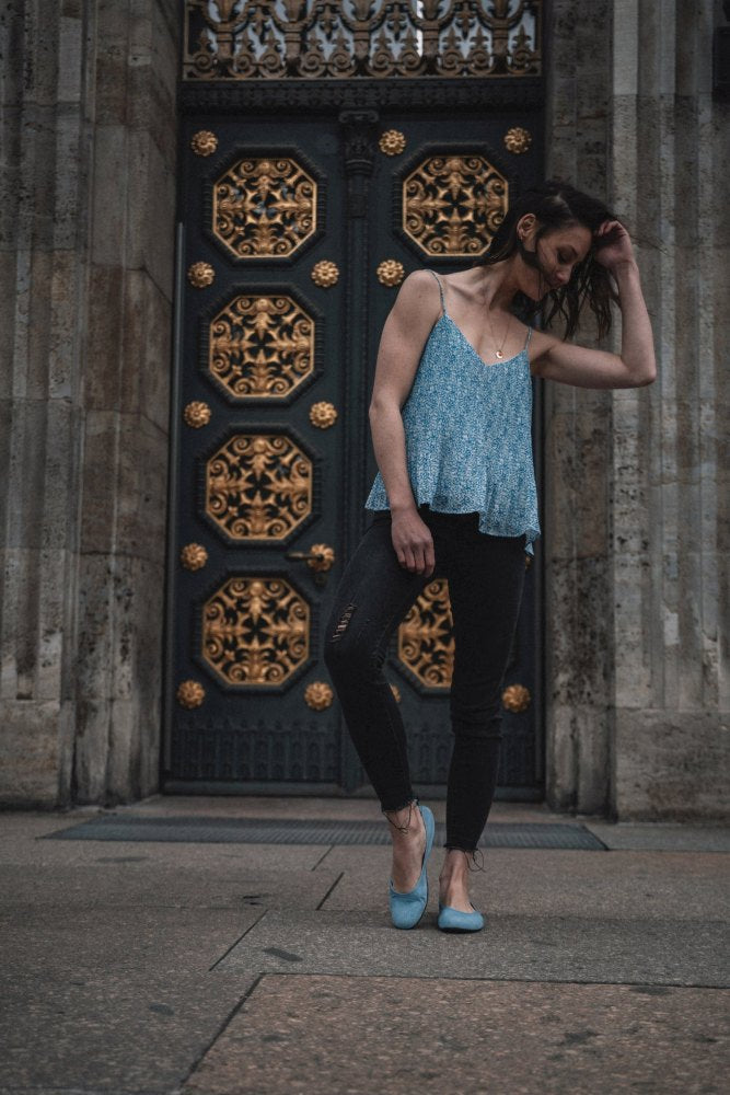 Een vrouw met lang haar staat voor een versierde deur met gouden details. Ze draagt het mouwloze TWIST Light Blue topje van ZAQQ, gecombineerd met zwarte jeans en flexibele schoenen. Ze kijkt naar beneden en raakt zachtjes haar haar aan in een omgeving die een elegante, sfeervolle atmosfeer uitstraalt.