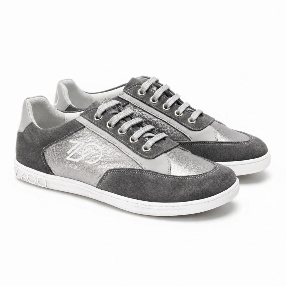 Een paar OQTAV Grey-sneakers van ZAQQ, gemaakt van fluweelzacht suède en gestructureerde inzetten. Met zilveren veters en het kenmerkende Zagoo-logo aan de zijkant en hiel worden deze stijlvolle schoenen naast elkaar op een witte achtergrond gepresenteerd.