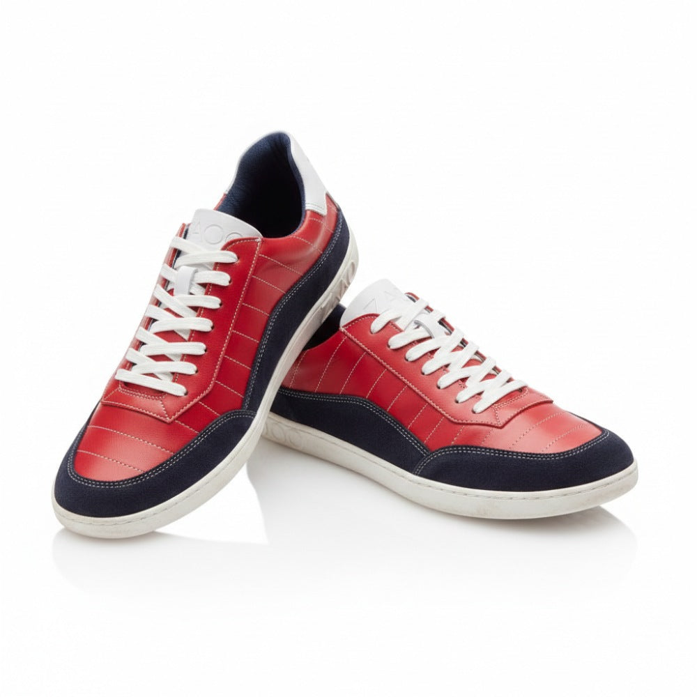 Wir präsentieren den QAMPION Navy Red von ZAQQ, ein Paar stylische Sneaker aus luxuriösem Nappaleder. Diese Sneaker zeichnen sich durch ein auffälliges rotes Obermaterial, weiße Schnürsenkel und marineblaue Akzente an Zehen und Sohlen aus. Kunstvoll auf einem weißen Hintergrund positioniert, lehnt sich ein Sneaker leicht an den anderen.