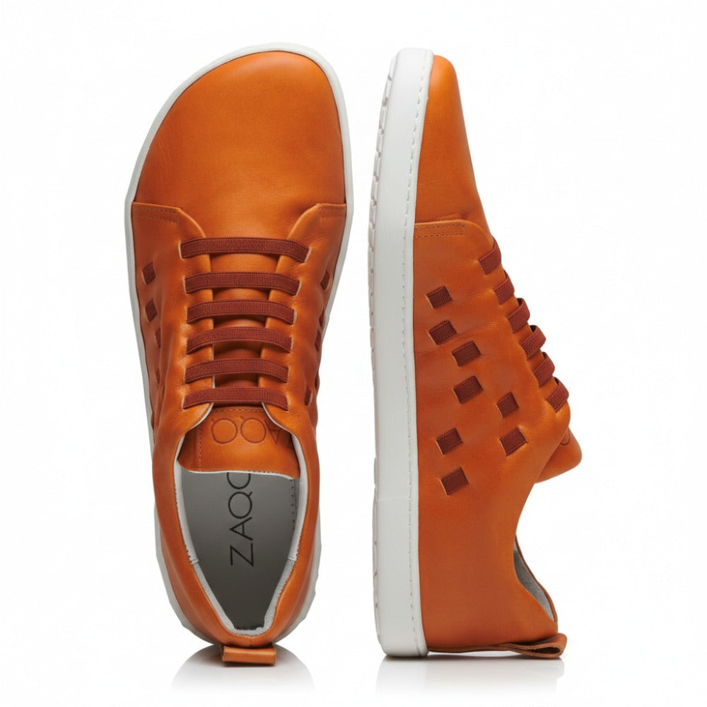 Die STRAQ Sierra-Sneaker von ZAQQ sind aus orangefarbenem Nappaleder gefertigt und haben weiße Sohlen. Sie zeichnen sich durch markante seitliche Aussparungen und ein Schnürdesign aus. Ein Sneaker wird aufrecht präsentiert, während der andere flach liegt, wodurch das ZAQQ-Branding sowohl auf der Innensohle als auch auf der Außenkante der Sohle hervorgehoben wird.