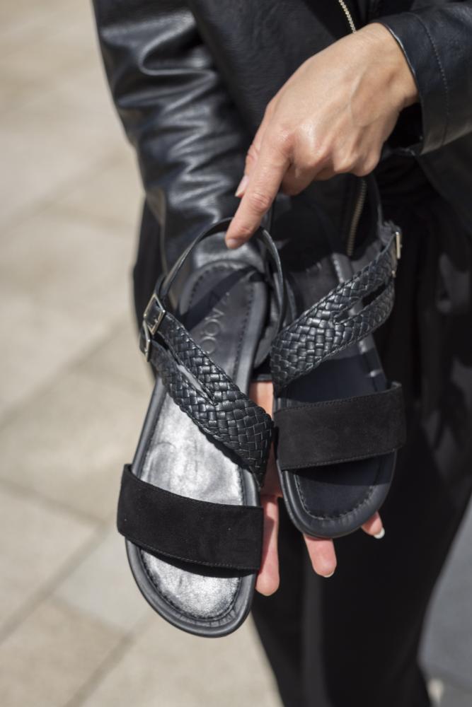 Die Person, bekleidet mit einer schwarzen Jacke und dunkler Hose, hält die Sandalen BROOQE Black von ZAQQ in der Hand. Diese Sandalen verfügen über gewebte Lederriemen aus Nappa- und Veloursleder. Die Zehenbänder aus Wildleder verstärken die feminine Eleganz der Sandalen auf dem gefliesten Außenboden.