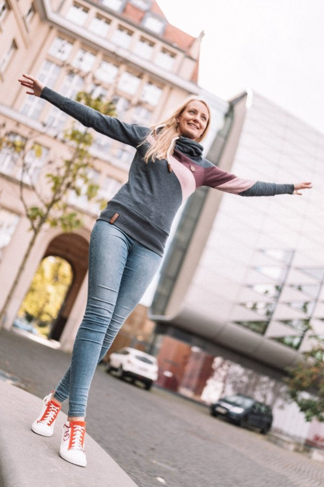 Eine Person mit langen Haaren balanciert mit ausgestreckten Armen auf einer niedrigen Mauer. Sie trägt einen bunten Pullover und blaue Jeans. An ihren Füßen trägt sie leuchtend rote ZAQQ CEDRIQ-Turnschuhe mit Kautschuksohle. Sie befindet sich in einer städtischen Umgebung mit modernen und traditionellen Gebäuden im Hintergrund.